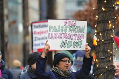 A woman holds a sign that reads 'Full Palestinian Liberation Now!' 
